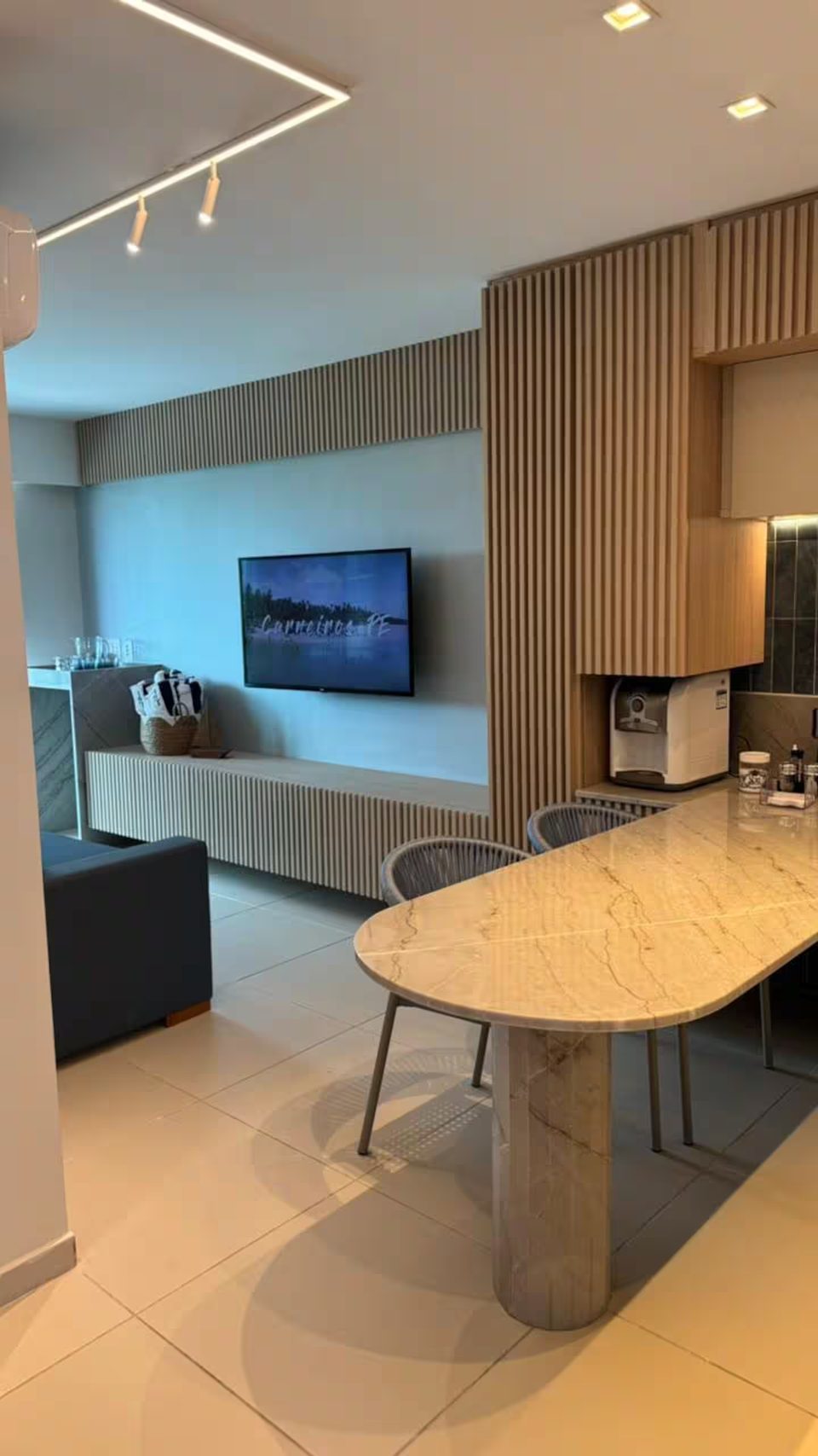 Sala de jantar do apartamento 302D com mesa de mármore, cadeiras, painel ripado de madeira e televisão.