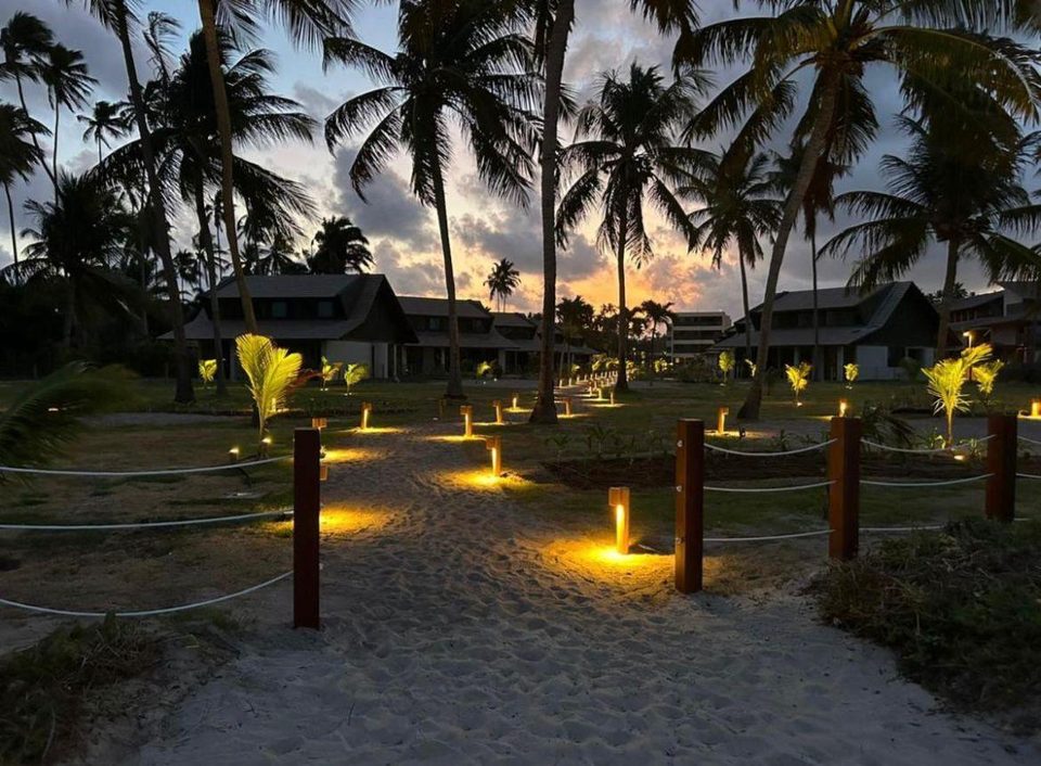 Acesso à praia iluminado ao anoitecer, entre coqueiros e a vegetação do condomínio.