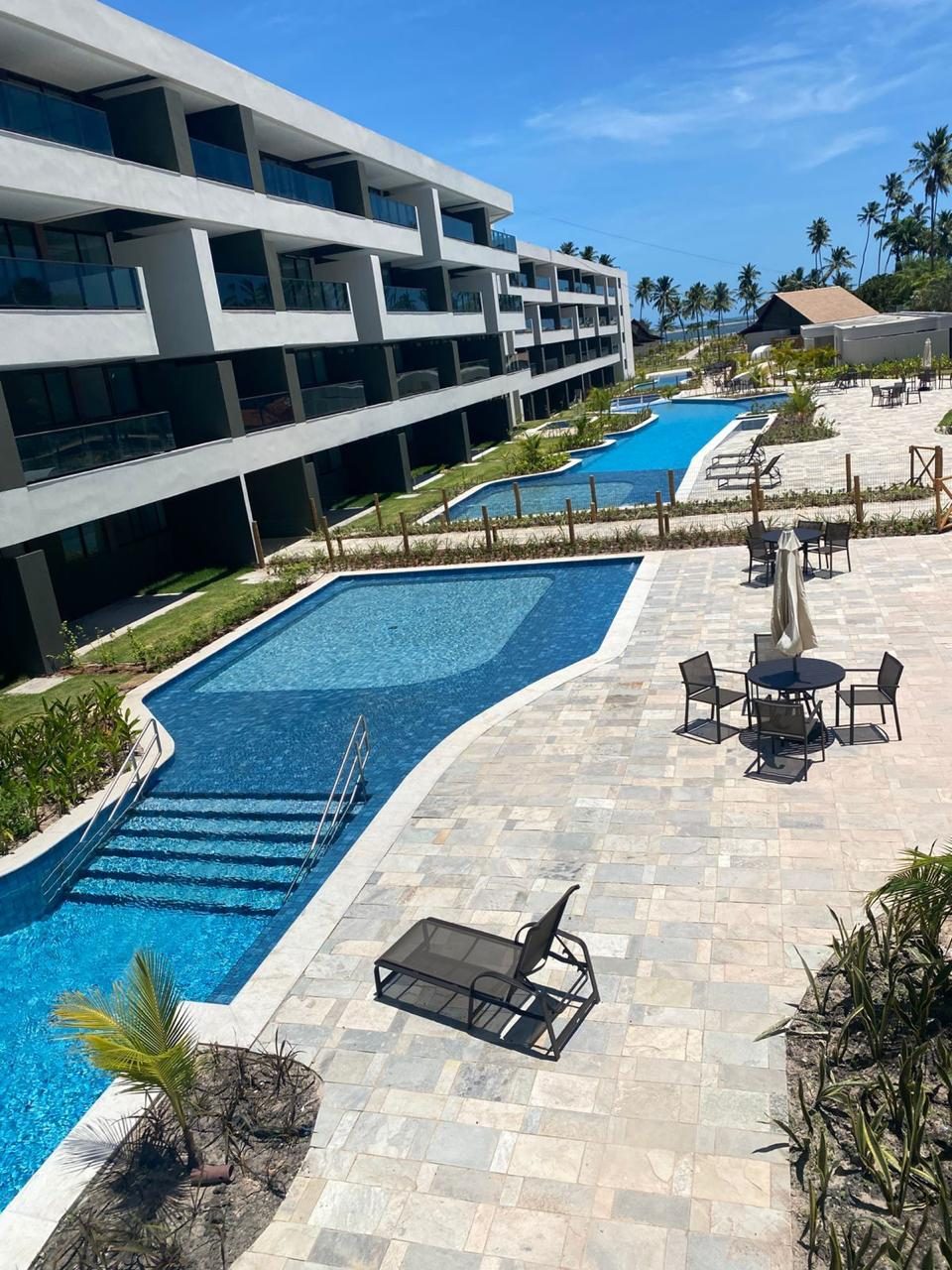 Piscina do condomínio com deck de pedra, espreguiçadeiras e vegetação ao redor.