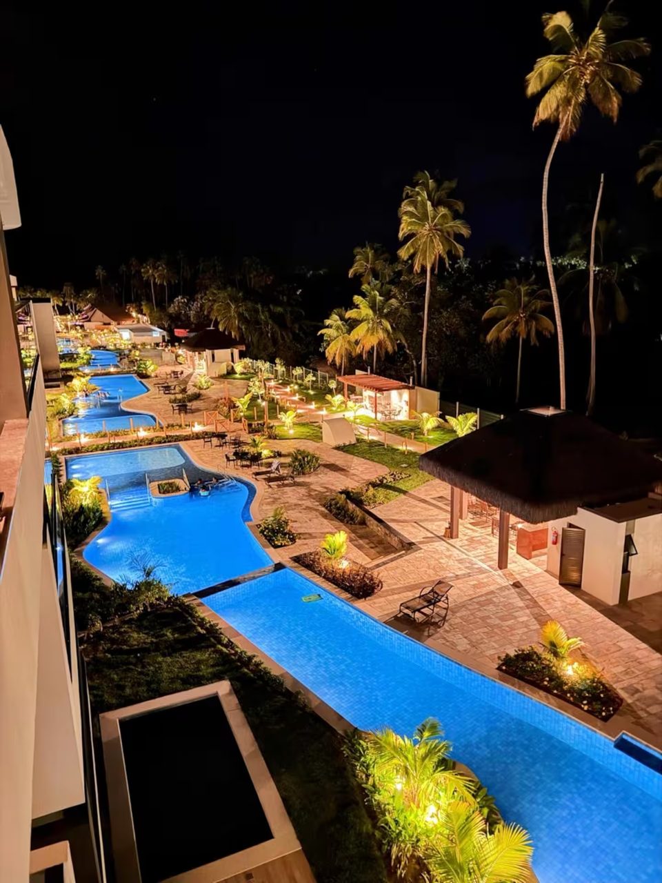 Piscinas do condomínio iluminadas à noite, vistas de cima, entre coqueiros.