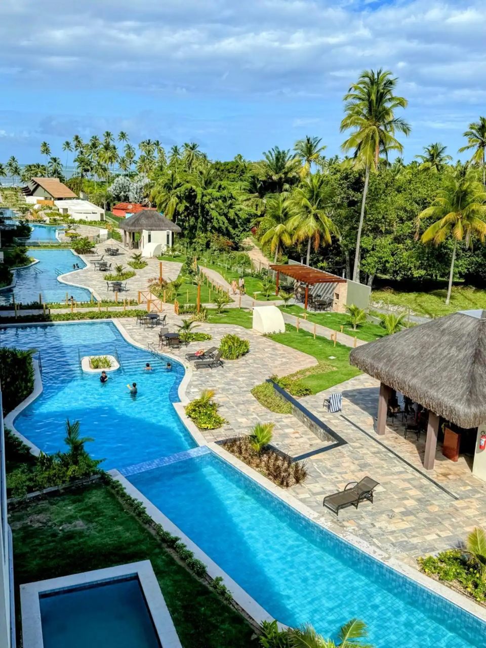 Vista das piscinas do Galápagos Beach Residence a partir da varanda: piscina em curva, coqueiros, bar de palha e o mar ao fundo.