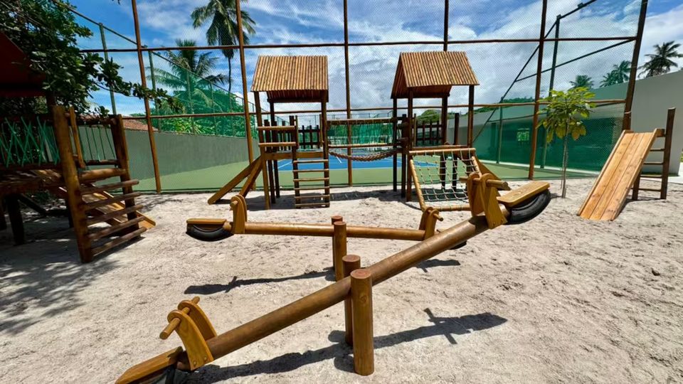 Playground de madeira do condomínio sobre areia, com escorregador e gangorra.